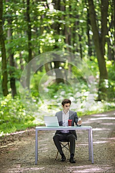 Businessman sitting at the office desk work at laptop computer and cup of coffee use mobile phone on road of green forest park. Bu