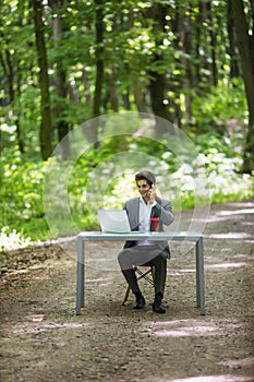 Businessman sitting at the office desk work at laptop computer and cup of coffee use mobile phone on road of green forest park. Bu