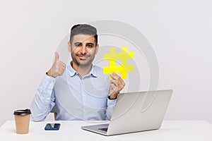 Businessman sitting in office workplace with laptop, holding big hashtag symbol and showing thumbs up, like gesture