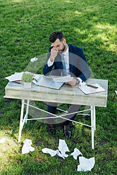 Businessman sitting in park behind table with laptop, smartphone, notebooks and flowerpot with many crumpled sheets of
