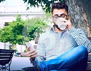 Businessman sitting on park bench with coffee using mobile phone.