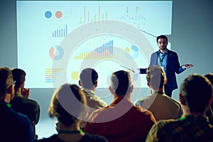 Businessman, speaker delivering presentation to attentive crowd with projection of colorful statistical graphs behind.