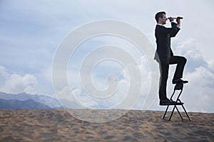 Businessman standing on a chair and looking through a telescope in the middle of the desert