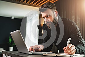 Businessman is standing by computer, looking at laptop screen, making notes in notebook. Man watching webinar, learning.