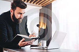 Businessman is standing by computer, looking at laptop screen, making notes in notebook. Man watching webinar, learning.