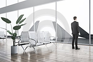 Businessman standing in creative airport waiting area interior with reflections on floor, wooden flooring, decorative plants,