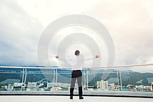 Businessman standing keeping arms raised on a roof and looking at city Success concept