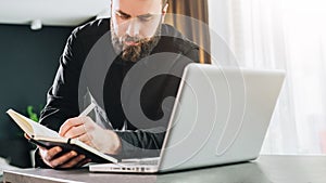 Businessman is standing near computer, working on laptop, making notes in notebook. Man watching webinar, learning.