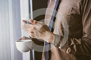 Businessman standing near window and using cell phone and holding coffee vintage tone