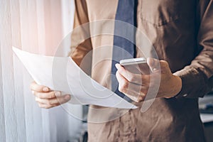 Businessman standing near window and using cell phone and holding paper sheet vintage tone