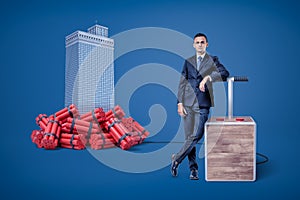 Businessman standing next to detonator box connected to heap of dynamite beside high-rise building in the background.