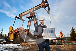 Businessman standing on an oilfield holding mini solar module next to an oil rig