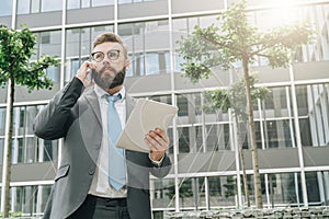 Businessman is standing outdoor,holding tablet computer and talking on his cell phone.Man working.Business conversation.