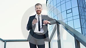 Businessman standing outside a modern corporate building with coffee cup and cellphone. Cityscape background