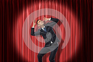 Businessman standing in spotlight near red stage curtain, leaning aside, looking up and protecting head with hands from