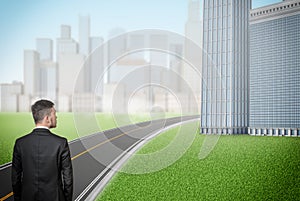 A businessman stands in front of a long empty road running between glass business buildings.