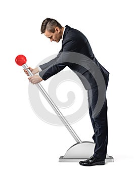 A businessman stands at a large lever with a red round knob and starts to move it.