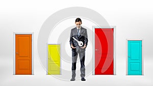 A businessman stands beside many small multicolored doors and looks at a large key hanging over his hands.