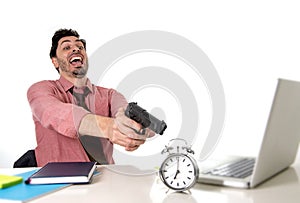 Businessman in stress at office computer desk pointing hand gun to alarm clock in out of time and project deadline expiring