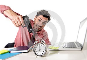Businessman in stress at office computer desk pointing hand gun to alarm clock in out of time and project deadline expiring
