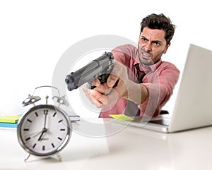 Businessman in stress at office computer desk pointing hand gun to alarm clock in out of time and project deadline expiring photo
