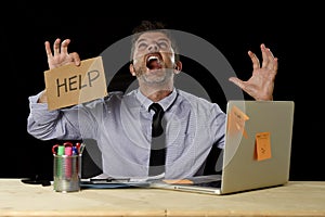 Businessman in stress working at office computer desk holding sign asking for help screaming crazy