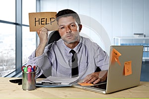 Businessman suffering stress working at computer desk holding sign asking for help looking tired exhausted