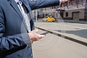 Businessman in a suit booking a taxi using mobile phone app standing on the street. yellow taxi car on the background of office