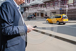 Businessman in a suit booking a taxi using mobile phone app standing on the street. yellow taxi car on the background of office