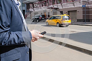 Businessman in a suit booking a taxi using mobile phone app standing on the street. yellow taxi car on the background of office