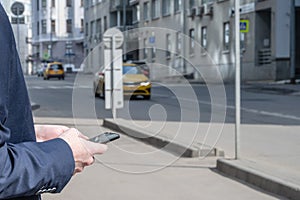 Businessman in a suit booking a taxi using mobile phone app standing on the street. yellow taxi car on the background of office