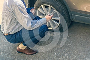 A businessman in suit crouching near his car with  punctured wheel. Man calling police using mobile phone. Street crime