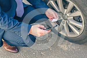 A businessman in suit crouching near his car with  punctured wheel. Man holding mobile phone and business card with data