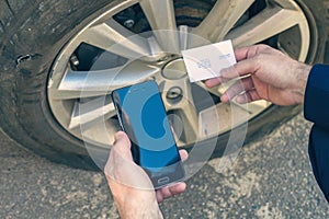 A businessman in suit crouching near his car with  punctured wheel. Man holding mobile phone and business card with data of isuran