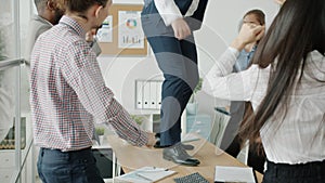 Businessman in suit dancing on table while multi-ethnic team clapping hands and partying indoors in office