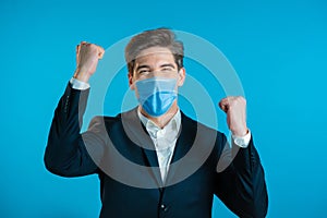 Businessman in suit and medical mask is very glad and happy, he shows yes gesture of victory, guy achieved result, goals