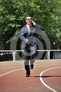 Businessman in suit and necktie running in stress on athletic track talking on mobile phone overworked