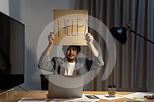 Businessman in suit sitting at office desk working on computer laptop asking for help holding cardboard sign