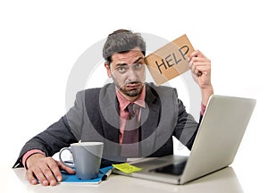 Businessman in suit and tie sitting at office desk working on computer laptop asking for help holding cardboard sign