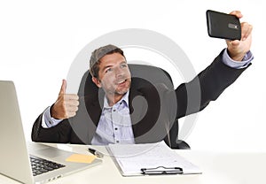 Businessman in suit working at office laptop computer desk using mobile phone for taking selfie photo