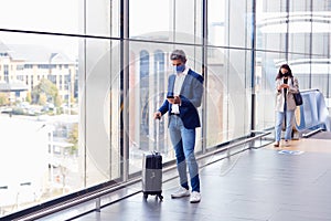 Businessman With Suitcase Standing By Window On Concourse At Railway Station With Mobile Phone