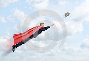 A businessman in a superhero cape flying in the sky trying to catch a 100 USD banknote.