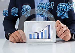 Businessman with tablet at desk with diagram of bar chart and cogs heads