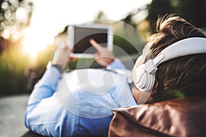 A businessman with tablet and headphones, lying on the bench outdoors at sunset.