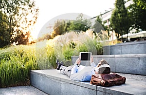 A businessman with tablet and headphones, lying on the steps outdoors at sunset.