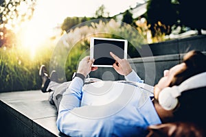 A businessman with tablet and headphones, lying on steps outdoors at sunset.