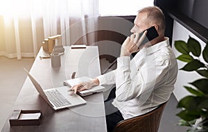 Businessman talking on mobile phone while looking at laptop screen. Business call in office