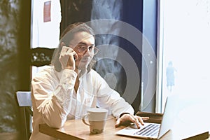 Businessman talking by mobile phone with opened laptop in cafe, co-working space. Having coffee-break