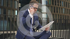 Businessman talking on phone, holding documents sitting on background of office building.