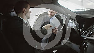 Businessman talking to seller and reading a contract while sitting in a car. Auto dealer is telling about details in a